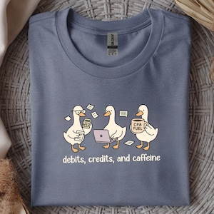 May include: A blue t-shirt with a cartoon design of three ducks. One duck uses a calculator, another a laptop, and the third holds a coffee mug. The text "debits, credits, and caffeine" is printed below the ducks.