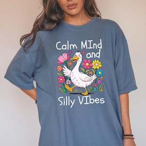 Puede incluir: Una camiseta azul con un ganso de dibujos animados blanco y flores. El texto "Calm Mind and Silly Vibes" está impreso en la camiseta.