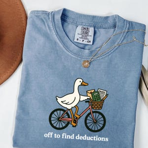 May include: A blue Comfort Colors t-shirt with a graphic of a white duck riding a pink bicycle, carrying tax-related items in a basket. The text "off to find deductions" is printed below the graphic. A gold necklace is draped across the shirt.