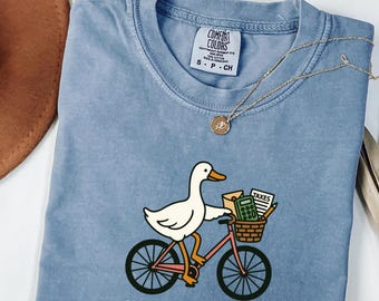 Könnte beinhalten: Blaues Comfort Colors T-Shirt mit einer Grafik einer weißen Ente auf einem rosa Fahrrad, die Steuerartikel in einem Korb transportiert. Der Text "off to find deductions" ist darunter gedruckt. Eine goldene Halskette liegt auf dem T-Shirt.
