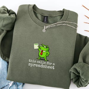 Può includere: Una felpa verde oliva con la scritta "this calls for a spreadsheet" e un ranocchio dei cartoni animati con occhiali da sole e cravatta rossa. Il ranocchio tiene in mano un foglio di calcolo. Una collana d'argento è drappeggiata sulla felpa.
