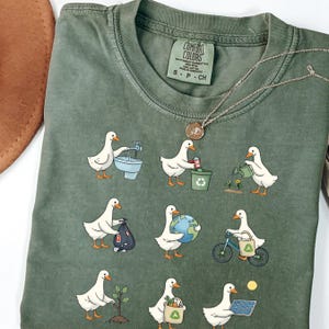 Puede incluir: Camiseta verde Comfort Colors con patos de dibujos animados realizando actividades ecológicas. Los patos se muestran cerrando un grifo, reciclando, regando plantas y montando en bicicleta. También se ve un collar plateado con un colgante.