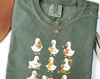 Può includere: T-shirt verde Comfort Colors con grafica di anatre che lavorano a maglia. Una collana d'argento con un piccolo ciondolo è drappeggiata sulla maglietta. La maglietta è piegata su una superficie bianca.