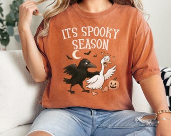 May include: Orange t-shirt featuring the text "IT'S SPOOKY SEASON" above a cartoon illustration of a black crow and a white goose, with a jack-o'-lantern and bats. The shirt is made of a soft material.