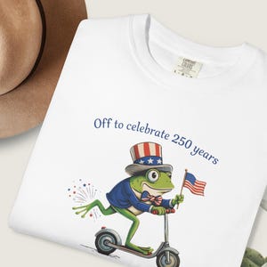 May include: White t-shirt with a cartoon frog riding a scooter and holding an American flag. The frog wears a patriotic hat and jacket. Text reads "Off to celebrate 250 years."