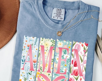 Puede incluir: Una camiseta azul Comfort Colors con la palabra "AMEN" en letras rosas, cada letra sobre un fondo floral. Un collar dorado con un colgante está drapeado sobre la camisa. La camisa tiene un pequeño lazo en la parte inferior.