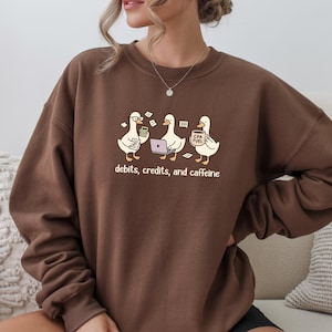 May include: A brown sweatshirt featuring a graphic of three cartoon ducks. One duck holds a calculator, another a laptop, and the third a coffee cup. The text "debits, credits, and caffeine" is below the ducks.