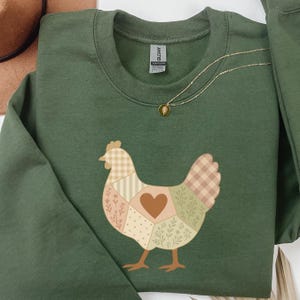 Op de afbeelding: Een groene sweatshirt met een patchwork kippenontwerp. De kip bestaat uit beige, bruine en groene patches, met een hart in het midden. Een gouden ketting met een kleine hanger is ook zichtbaar.
