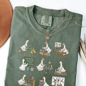 May include: A sage green Comfort Colors t-shirt with a science-themed illustration. The design shows cartoon geese in lab coats performing experiments. The text "SCIENCE EXPERIMENT" and other science-related phrases are also visible. A silver necklace with a pendant is draped across the shirt.