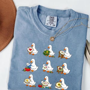 May include: A pale blue Comfort Colors t-shirt with a pattern of white ducks embroidering. The ducks are shown with various embroidery projects. A gold necklace and a brown hat are also visible.