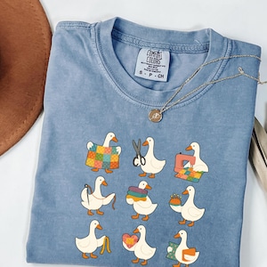 May include: A light blue Comfort Colors t-shirt featuring a whimsical design of cartoon ducks engaged in various sewing and quilting activities. The ducks are depicted with scissors, sewing machines, and colorful fabric. A gold necklace with a pendant is draped across the shirt.