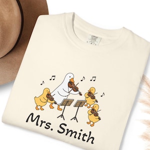 Puede incluir: Una camiseta de color crema con un gráfico de un pato grande blanco y tres patitos amarillos tocando el violín frente a atriles. Notas musicales flotan sobre ellos y el texto "Mrs. Smith" está impreso en negro debajo del gráfico.