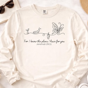 May include: A cream-coloured long-sleeve shirt featuring a black line-art graphic of a life cycle from sprout to caterpillar, chrysalis, and butterfly. Below the graphic, the text reads: For I know the plans I have for you, Jeremiah 29:11.