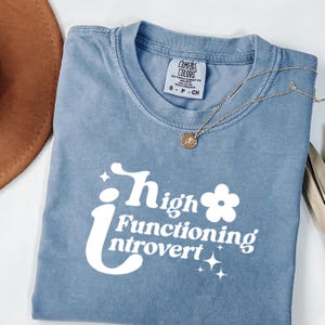 Könnte beinhalten: Ein blaues Comfort Colors T-Shirt mit den Worten "High Functioning Introvert" in Weiß. Das Shirt hat einen Rundhalsausschnitt und kurze Ärmel. Eine goldene Halskette mit Anhänger liegt über dem Shirt.
