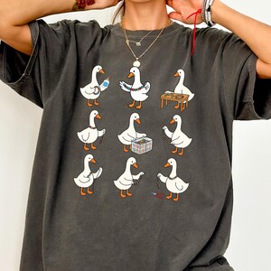 May include: Dark gray t-shirt featuring a whimsical design of white geese crafting jewelry. Each goose is depicted with orange feet and beaks, holding beads, tools, and finished necklaces. The design is repeated in a grid pattern across the shirt.
