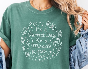 May include: A teal green t-shirt with the text "It's a Perfect Day for a Miracle" in white, surrounded by line art of hearts, flowers, a syringe, footprints, and a thermometer. The shirt has short sleeves.