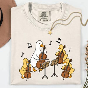 May include: A cream-colored t-shirt featuring a graphic of one white duck and three yellow ducklings playing cellos. The shirt has a Comfort Colors label at the neck, size XL-XG-TG, and is accompanied by a gold star necklace.