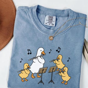 May include: A blue t-shirt featuring a graphic of a white adult duck and three yellow ducklings playing silver flutes. The ducks stand before music stands with black musical notes floating around them. The shirt has a Comfort Colors brand label.