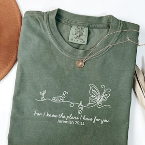 以下が含まれることがあります： オリーブグリーンのComfort Colors Tシャツ。白いイモムシ、サナギ、蝶のグラフィック。テキスト「For I know the plans I have for you Jeremiah 29:11」が下に印刷されています。ペンダント付きのゴールドネックレスがシャツの上にあります。