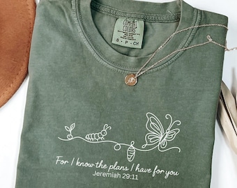 May include: Olive green Comfort Colors t-shirt with white graphic of a caterpillar, chrysalis, and butterfly. The text "For I know the plans I have for you Jeremiah 29:11" is printed below the graphic. A gold necklace with a pendant rests on the shirt.