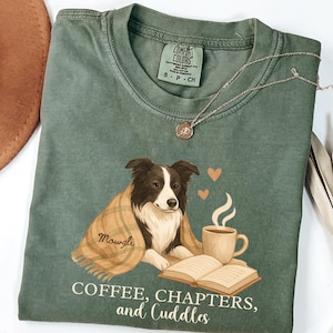 May include: Green t-shirt with a graphic of a dog wrapped in a blanket, a coffee cup, and an open book. The text "COFFEE, CHAPTERS, and CUDDLES" is printed below the graphic. A silver necklace is draped across the shirt.