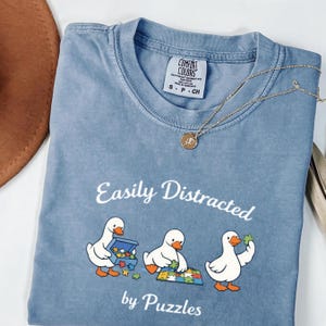 Puede incluir: Una camiseta azul Comfort Colors con la frase "Easily Distracted by Puzzles" y un gráfico de tres patos de dibujos animados armando rompecabezas. Un collar dorado con un colgante está sobre la camiseta.