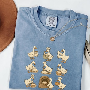May include: A light blue Comfort Colors t-shirt featuring a whimsical design of cartoon geese baking bread. The geese are depicted in various baking activities, such as mixing ingredients, kneading dough, and carrying bread. A gold necklace with a pendant is draped across the shirt.