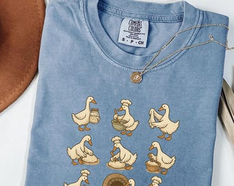 May include: A light blue Comfort Colors t-shirt featuring a whimsical design of cartoon geese baking bread. The geese are depicted in various baking activities, such as mixing ingredients and carrying loaves. A gold necklace with a pendant is also visible.