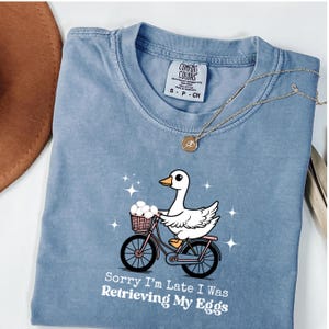 Op de afbeelding: Lichtblauw T-shirt met een cartoon gans op een fiets met eieren. De tekst "Sorry I'm Late I Was Retrieving My Eggs" staat eronder. Een gouden ketting ligt op het shirt. Comfort Colors label.