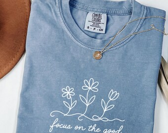 May include: A blue Comfort Colors t-shirt with a white floral design and the phrase "focus on the good." A gold necklace with a small pendant rests on the shirt. The shirt is folded and has a tag at the neckline.