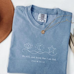 Könnte beinhalten: Ein blaues Comfort Colors T-Shirt mit dem Text "Be still, and know that I am God. PSALM 46:10" und einer weißen Grafik einer Lotusblume, eines Mondes, einer Sonne und eines Sterns. Eine goldene Halskette mit Anhänger liegt auf dem Shirt.