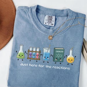 Pode incluir: Uma t-shirt azul com um gráfico de cinco personagens de química sorridentes, incluindo gobelés, tubos de ensaio e um livro com a etiqueta Chemistry. Abaixo das personagens, o texto diz Just here for the reactions. Etiqueta Comfort Colors.