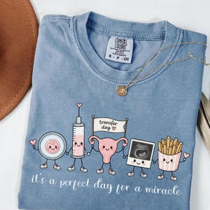 Puede incluir: Una camiseta azul con un gráfico de cinco personajes de dibujos animados: una célula, una jeringuilla, un útero con un cartel que dice transfer day, una ecografía y patatas fritas. El texto dice it's a perfect day for a miracle.
