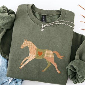 May include: An olive green sweatshirt featuring a patchwork horse design with plaid, floral, and heart details. The horse is in shades of tan, green, and brown. A silver necklace with a sunburst pendant is draped across the sweatshirt.