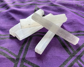 May include: Four white selenite wands, also known as selenite sticks, are stacked on top of each other on a purple fabric background. The wands are smooth and have a natural, crystalline texture.