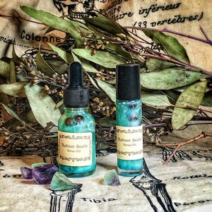 May include: Two glass bottles of "Radiant Health Ritual Oil" with teal liquid and a label. One bottle has a dropper, the other a rollerball. Amethyst and fluorite crystals are scattered around the bottles, with dried leaves in the background.