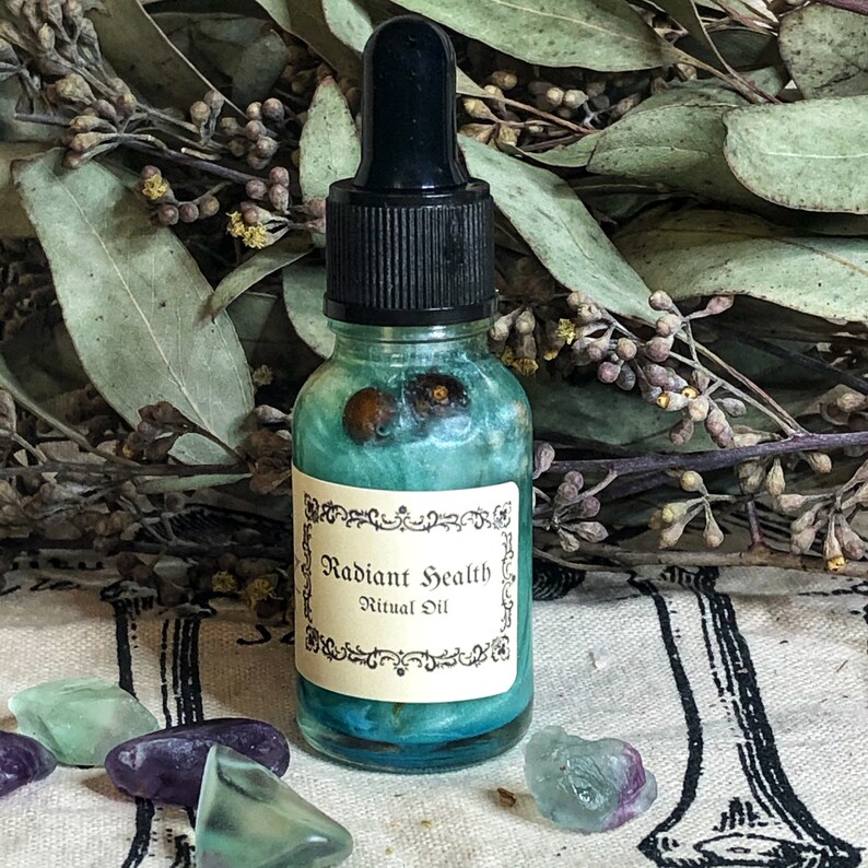May include: A small glass bottle with a black dropper top, filled with a teal liquid and decorative elements. The label reads "Radiant Health Ritual Oil" in an ornate font. The bottle is surrounded by crystals and dried foliage.