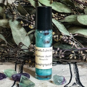 May include: A glass roll-on bottle with a black cap, filled with a teal liquid. The label reads "Radiant Health Ritual Oil". The background features dried leaves and colourful crystals. The bottle is approximately 10 cm tall.