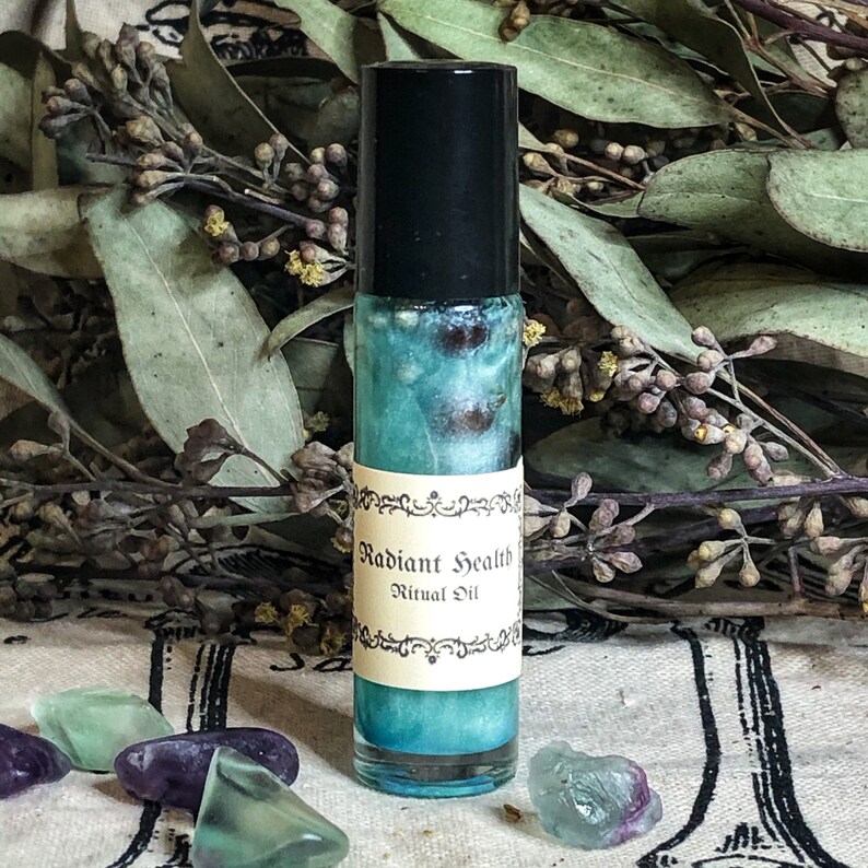 May include: A glass roll-on bottle with a black cap, filled with a teal liquid. The label reads "Radiant Health Ritual Oil". The background features dried leaves and colourful crystals. The bottle is approximately 10 cm tall.
