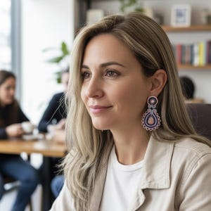 Puede incluir: Una mujer lleva pendientes grandes y ornamentados. Los pendientes tienen forma de lágrima con un diseño azul oscuro y rosa, y un borde festoneado blanco. Lleva una camiseta blanca y una chaqueta beige claro. El fondo muestra un entorno de oficina borroso.