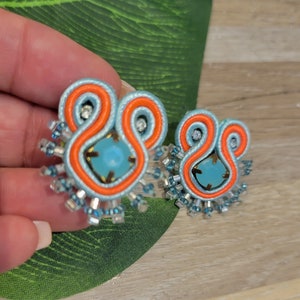 Puede incluir: Un par de pendientes hechos a mano con una piedra central azul claro, rodeada de bordados naranja y azul claro. Los pendientes están adornados con pequeñas cuentas claras y azules, creando un diseño artístico único.