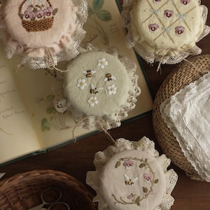 Könnte beinhalten: Eine Sammlung von bestickten Stoffkreisen mit Blumen- und Bienenmotiven. Jeder Kreis ist mit Spitze verziert und weist handgenähte Details auf, darunter ein Blumenkorb, Stiefmütterchen und Bienen. Die Farbpalette umfasst zarte Rosa-, Gelb- und Grüntöne.