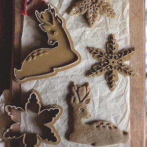 Peut inclure: Vue aérienne de biscuits et emporte-pièces de Noël sur du papier sulfurisé. Les biscuits ont la forme de flocons de neige et de cerfs. Un petit rouleau à pâtisserie et des cloches dorées sont également présents, avec un ruban de velours rouge.