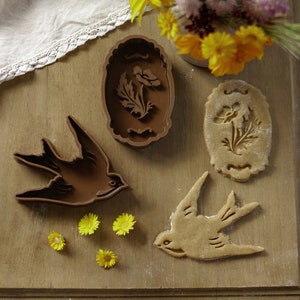 May include: A brown swallow-shaped cookie cutter and a baked cookie in the same shape, alongside a brown floral-shaped cookie cutter and a baked cookie. Yellow flowers and a bouquet of flowers are also present on a wooden surface.
