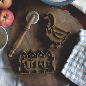 以下が含まれることがあります： ガチョウをテーマにしたパイ皿、クッキーカッター、ペストリーホイールなど、ベーキングツールとアクセサリーのコレクション。お皿には赤いリボンをつけたガチョウが描かれています。カッターはガチョウとフェンスの形をしています。リンゴとスパイスのボウルも写っています。