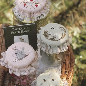 May include: Three glass jars with fabric covers featuring embroidered designs of radishes, a watering can, and bees with flowers. Each cover is trimmed with white lace. A book titled The Tale of Peter Rabbit is placed behind the jars.