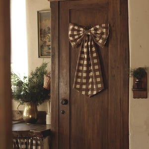 May include: A large decorative bow made of brown and white gingham fabric attached to the front of a tall, dark wooden cabinet door. The bow features two wide loops and two long, pointed tails hanging vertically down the center of the door panel.