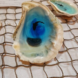 May include: A blue and white oyster shell with a black silhouette of a fish inside. The shell is sitting on a brown net-like surface.