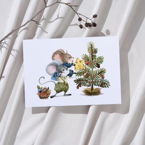 Puede incluir: Ilustración en acuarela de tres ratones decorando un pequeño árbol de Navidad. Un ratón sostiene un adorno de queso en forma de estrella. El árbol está adornado con bayas y bellotas. Una pequeña cesta de arándanos está cerca. La imagen está en una tarjeta blanca.