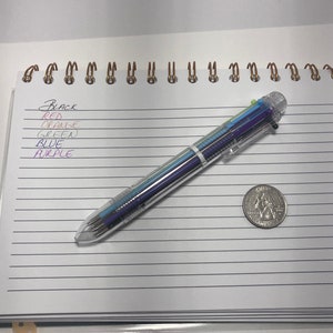 May include: A clear multi-colored pen with black, red, orange, green, blue, and purple ink. The pen is lying on a white lined notebook with a US quarter coin next to it.